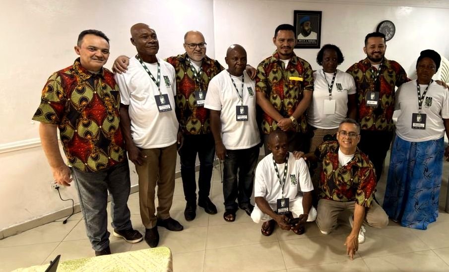 Irmãos nigerianos e visitantes brasileiros, alguns com camisas de estampa africana, reunidos para uma foto no salão.