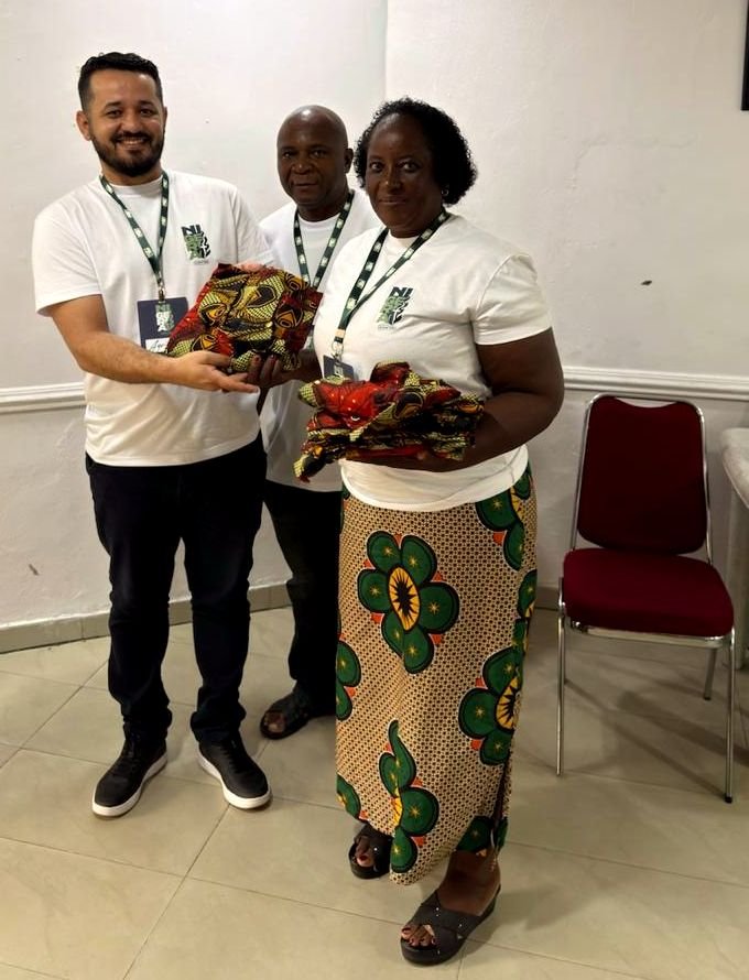 Um visitante brasileiro e uma irmã nigeriana seguram tecidos estampados trocados como presente, ao lado de um irmão nigeriano.