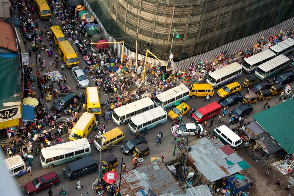 Vista do alto de uma rua movimentada de Lagos, com ônibus amarelos, vans e muita gente entre as bancas de comércio.